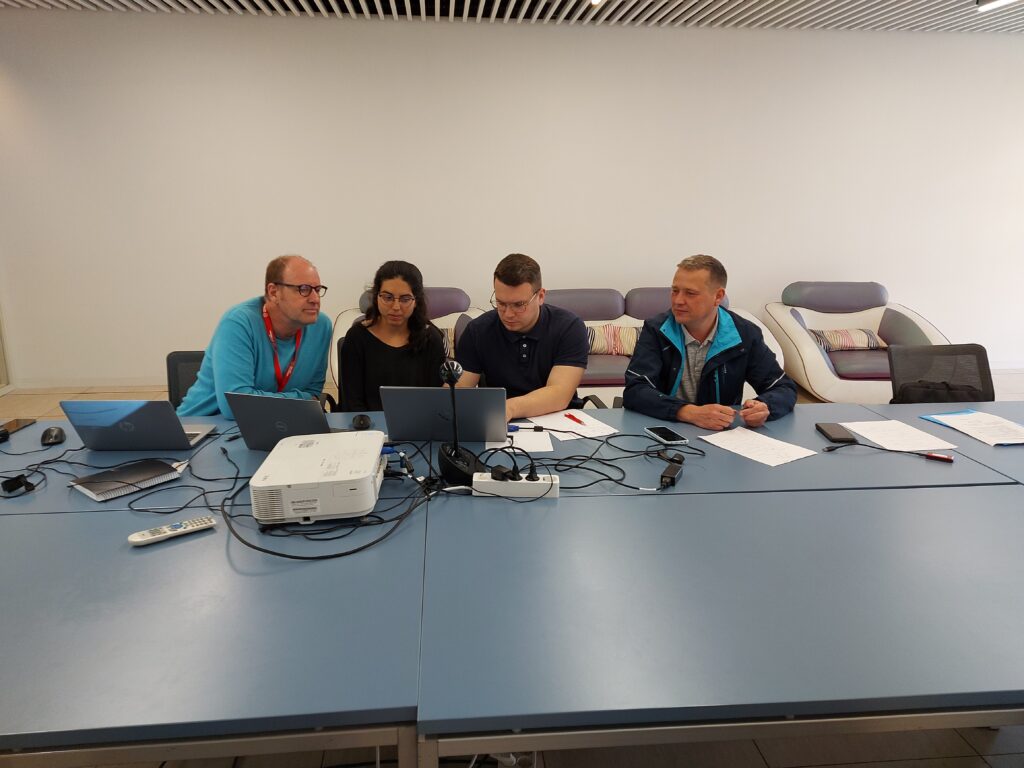 Image shows 3 men and one women in a conference room, in front of three laptops, a projector, and paper notes.