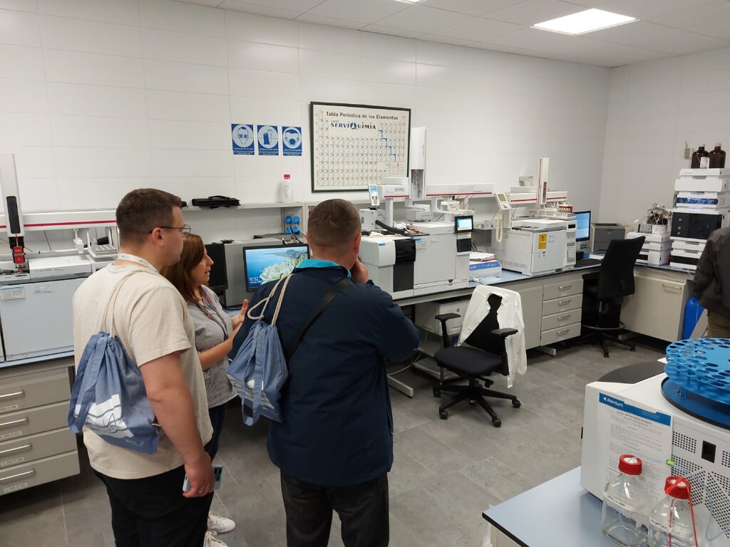 The image shows Josepa Fabregas (CAT), Serhii Martynov and his colleague (NUWEE) in a water quality monitoring lab in L'Ampolla DWTP.