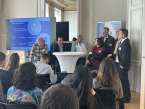 The picture shows a panel discussion of 7 people at a round table. In the background on the left a screen displaying the speakers of the SafeCREW Final Conference panel discussion (dark and light blue, inclduing EU logo). A roll up with SafeCREW logo behind the speakers. Audience in the foreground. Hanse Office conference room in art nouveau style with large mirror in the back. (C) M. Remmert-Rieper, Tutech Innovation GmbH.